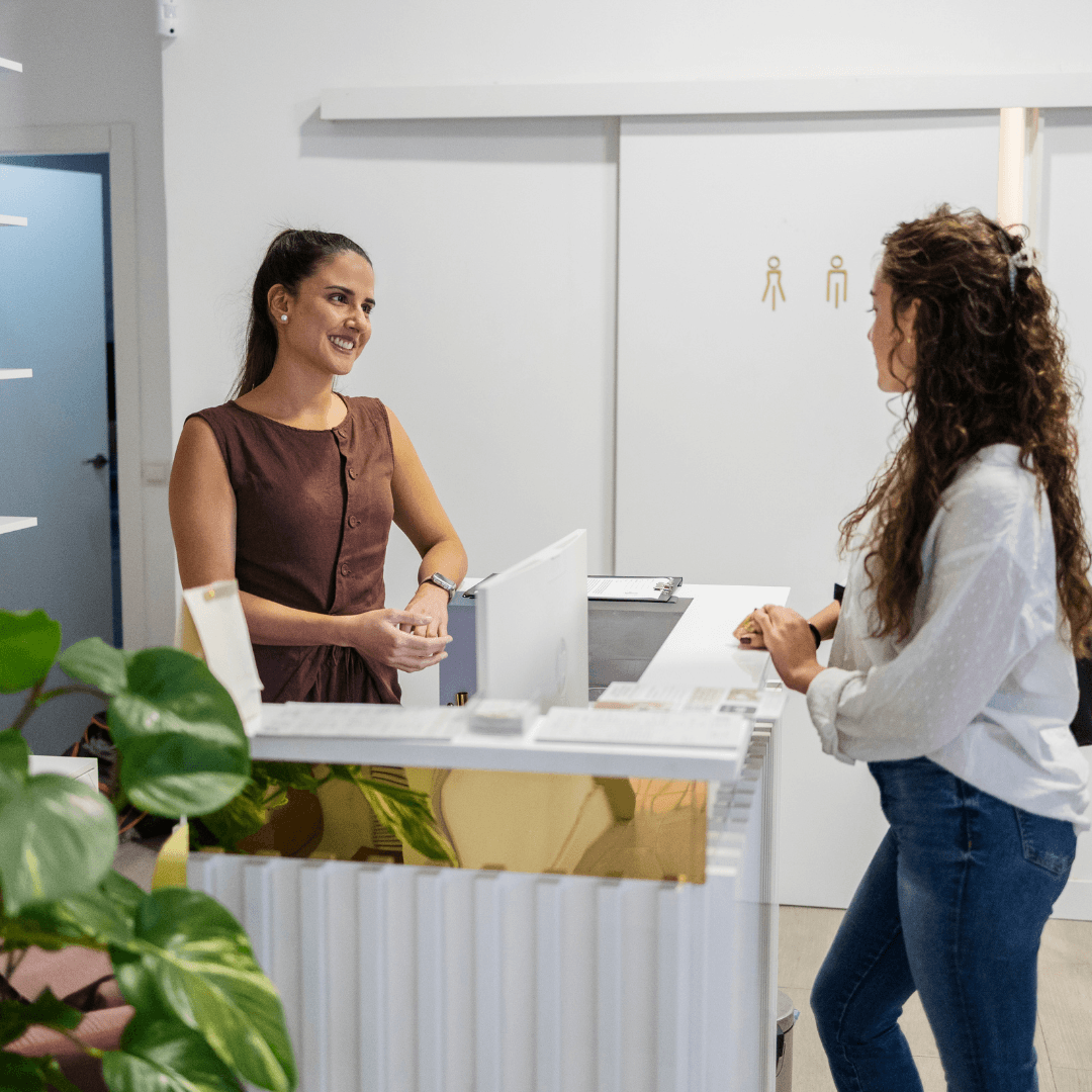 Two women speaking at a clinic reception desk in a professional healthcare setting